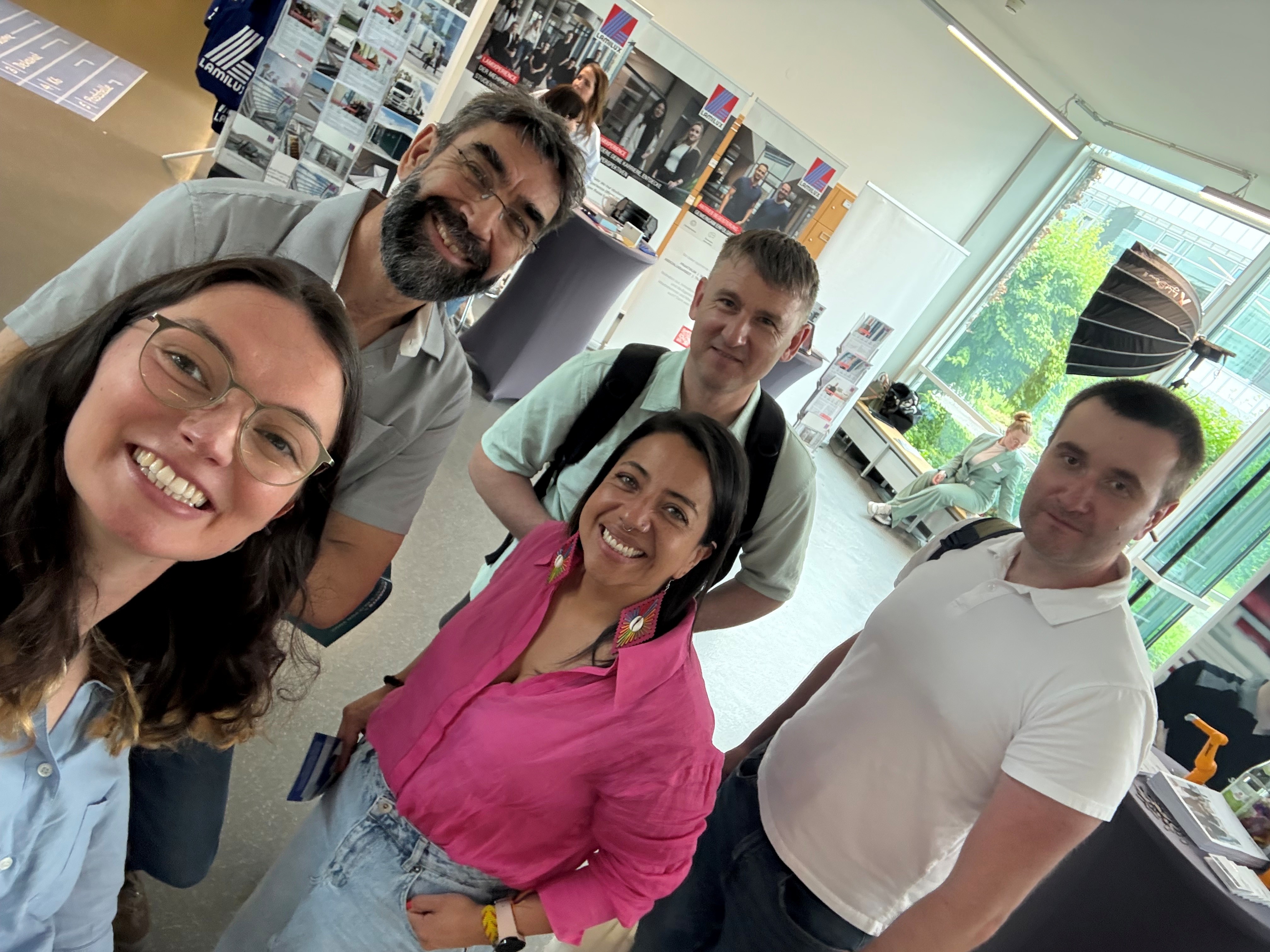 Ein Selfie einer Gruppe mit fünf Personen. Alle lächeln in die Kamera. Sie befinden sich auf einer Jobmesse, im Hintergrund sind Messerstände zu sehen.